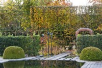 Jardin contemporain avec étang et topiaires de buis, séparé du salon de jardin voisin par des haies de houx, des sculptures ornementales en métal et un accès en pierre à l'espace de détente, adossé à une haute haie de charmes.