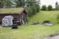 Abri en bois au toit d'herbe près de l'entrée du jardin botanique de Tromsø. Petites plantes en pot dans un parterre de fleurs surélevé dans l'herbe. Outils de jardin et sacs de compost. Au milieu de l'été.