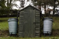 Un petit cabanon situé dans un jardin boisé avec des réservoirs d'eau de chaque côté. Whitstone Farm, Devon NGS jardin, automne