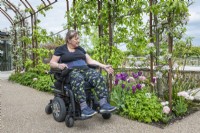 Patient en fauteuil roulant sous l'arche en acier rouillé. Les plantes comprennent : des pommiers en fleurs formés avec des tulipes 'Angelique' et 'Recreado'.Horatio's Garden South West - SalisburyThe Duke of Cornwall Spinal Treatment Center