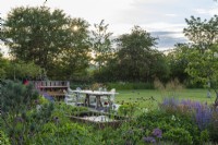Terrasse bordée de parterres plantés de scabieuses, d'avoine dorée, d'herbes à chats, d'alliums et de geums. Dispositif d'eau de réservoir en acier rouillé.