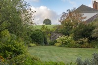 Vue sur la pelouse, devant les hortensias 'Annabelle' et les hakonechloa dorés, jusqu'à une porte menant à un jardin de devant clos. Derrière, une colline s'élève brusquement.