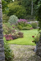 Vue à travers une vieille porte en pierre dans le jardin du ruisseau avec un parterre de fleurs sinueux planté de plantes vivaces herbacées et de graminées ornementales.