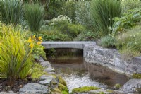 Le ruisseau coule sous un pont et dans le jardin boisé planté d'iris, de géraniums vivaces, de saules et de bullwort.