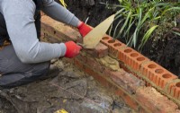 Un ouvrier utilisant une truelle de maçonnerie pour construire un muret de briques autour d'un parterre de fleurs surélevé lors de la rénovation d'un petit jardin londonien.