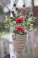 Bouquet d'hiver comprenant une rose de Gueldre et du gui avec des baies, des brindilles de mélèze avec des cônes et du feuillage d'ilex dans un pot en corde suspendu fait maison.
