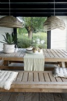 Table et bancs en bois dans le pavillon avec lampes et plantes et vue sur le saule et les herbes.