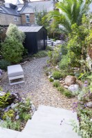 Petit jardin avec cabanon noir et chemin de gravier incurvé qui traverse le milieu vers un coin salon