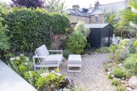 Petit jardin avec cabanon noir et chemin de gravier incurvé qui traverse le milieu vers un coin salon avec chaise et repose-pieds au premier plan