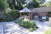 Jardin avec terrasse carrelée en pierre, allée carrelée en quinconce, gazebo rond en bois et parterre de fleurs au milieu avec plantation mixte