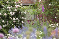 Dépendance rustique en brique avec Rosa 'Généreux Jardinier' vue à travers les parterres de fleurs et la pelouse du pré