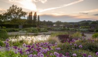 Vue du lever du soleil sur le jardin de gravier en direction du lac Blagdon - diverses variétés d'alliums et parterres de fleurs mixtes et petit étang
