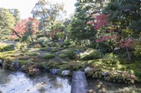 Pont en pierre traversant le ruisseau avec vue sur les monticules d'herbe ondulés dans le jardin, les arbres et arbustes aux couleurs d'automne.