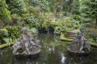 Deux statues figuratives dans un étang, avec de petites cascades en arrière-plan, des marches à droite et une végétation tropicale luxuriante en arrière-plan. Jardins du Monte Palace, Madère 
