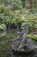 Une statue figurative dans un étang, avec de petites cascades en arrière-plan, des marches à droite et une plantation tropicale luxuriante en arrière-plan. Jardins du Monte Palace, Madère 