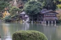 Vue sur l'étang Kasumigaike jusqu'au bâtiment de la Maison de Thé appelé Uchihashitei et sur plusieurs arbres avec des wigwams de bambou et de corde comme protection contre les dommages causés par la neige appelés Yukitsuri. 