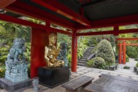 Un grand Bouddha doré assis dans une pagode rouge de style oriental avec des statues de chien Foo, des meubles en bois et un jardin tropical en arrière-plan. Jardin du Monte Palace, Madère. Août. 