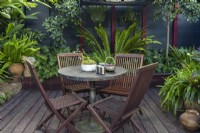 Un pot de plantes grasses mélangées, une théière et des tasses sur une table en bois avec des chaises, dans un jardin subtropical. 
