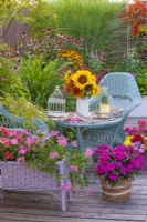 Parterre de fleurs surélevé planté de pélargoniums, un pot en osier avec des Impatiens, un bouquet de fleurs d'été dont des tournesols et des meubles en osier sur une terrasse en bois. 