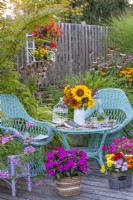 Parterre de fleurs surélevé planté de pélargoniums, un pot en osier avec des Impatiens, un bouquet de fleurs d'été dont des tournesols et des meubles en osier sur une terrasse en bois. 