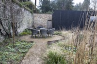 Un chemin ébréché mène à une section circulaire avec une table ronde en rotin et des chaises. Diverses herbes et plantes se trouvent à droite. Hiver. Jardin italien à Great Ambrook. 