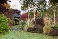 Urne en terre cuite entourée d'arbustes, de graminées ornementales et d'arbres dont Prunus serrula et bouleaux à tige blanche en novembre 