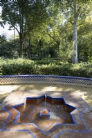 Une fontaine basse et géométrique avec des carreaux vernissés colorés de style mudéjar et islamique, avec une variété de feuillages et un banc carrelé bas et incurvé en arrière-plan. Parque de Maria Luisa, Séville, Espagne. Septembre 
