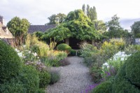 Un chemin de gravier mène au-delà des topiaires et des parterres de fleurs jusqu'à une maison d'été couverte de glycines dans le jardin fleuri du Manoir, Little Compton. 