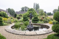Un étang circulaire avec fontaine est entouré d'un chemin de gravier et de parterres de fleurs avec des topiaires et des plantes vivaces dans le jardin fleuri du Manoir, Little Compton. 