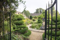 Une vue à travers les portes du jardin fleuri avec des plantes vivaces, des topiaires et un étang circulaire au Manoir, Little Compton. 