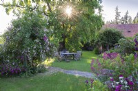 Jardin campagnard avec grande pelouse avec salon de jardin en bois sous un arbre mature. À gauche se trouve un rosier grimpant et à droite un parterre mixte de plantes vivaces. 