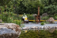 Une piscine avec bord de galets et gros rocher menant à un coin salon avec poêle à bois extérieur - Hurtigruten : The Relation-Ship Garden - designer Max Parker-Smith - RHS Hampton Court Flower Palace Garden Festival 2023. 