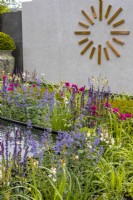 Un parterre de fleurs vivaces colorées planté de Nepeta, Salvia et Ixia 'Mabel'. Une grande horloge murale moderne en cuivre décore un mur carrelé en arrière-plan. Juin. Designer : Kevin Dennis, Bord Bia Bloom 2023 