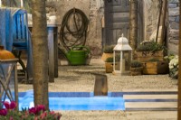 Cour italienne contemporaine devant la maison en pierre avec canal d'eau coulant sur la surface de gravier dirigé vers un petit étang avec des bougies flottantes. Présentation d'herbes aromatiques en pots et lanterne.  