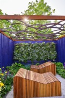 Espace détente entouré de panneaux peints bleu marine avec transats décoratifs en bois adossé à un mur végétal vivant sous un toit de pergola ajouré au motif géométrique. JuinBord Bia Bloom, DublinDesigner : Jane McCorkell 