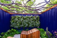 Espace détente entouré de panneaux peints bleu marine avec transats décoratifs en bois adossé à un mur végétal vivant sous un toit de pergola ajouré au motif géométrique. JuinBord Bia Bloom, DublinDesigner : Jane McCorkell 